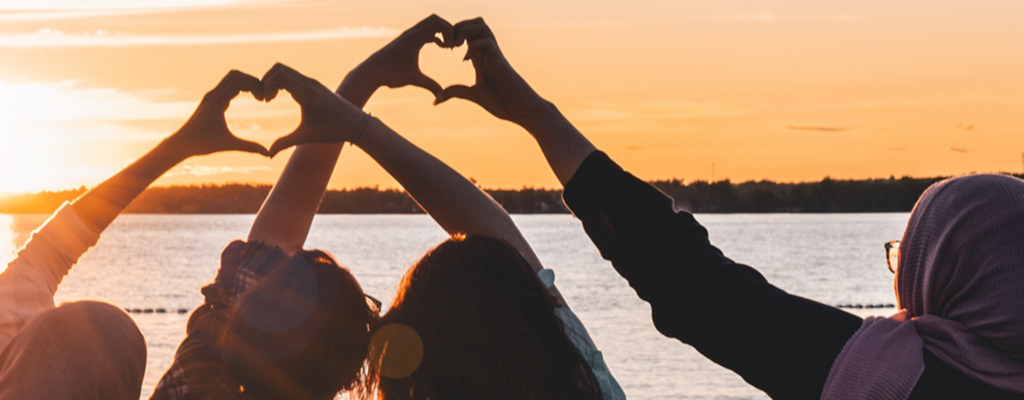 Four people making heart silhouettes with their hands against a sunset backdrop